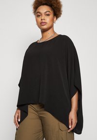 Black poncho-style top featuring a loose, flowing design, textured fabric, and wide arm openings, paired with khaki cargo pants.