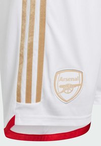 White athletic shorts with gold stripes, red trim, and an embroidered club logo featuring a cannon on a shield.