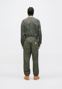 Zielona bluza w kamuflażu z długim rękawem oraz oliwkowe spodnie z dwoma tylnymi kieszeniami, wykonane z trwałej tkaniny. Prezentują solidne brązowe buty robocze.