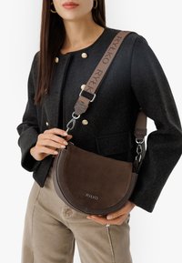 Braune Wildleder- und Leder-Crossbody-Tasche mit geschwungener Form, ausgestattet mit einem markierten Schulterriemen und silbernen Hardware-Details.
