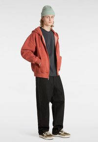 Orange zip-up hoodie i bomullsblandning med två framfickor, matchad med en mörkgrå t-shirt, svarta löst sittande byxor och grön stickad mössa.