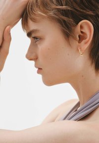 Pendientes de aro dorados con un acento de estrella, que exhiben una textura suave y un diseño circular. Llevados junto a un escote de tela gris suave.