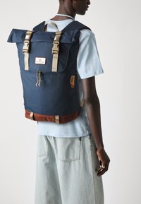 Marine roll-top ryggsekk laget av slitesterkt stoff, med brune stropper, brun bunn og en logo-patch. Praktisk design med glidelås og lommer.