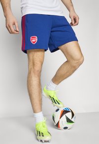 Mėlyni sportiniai šortai su raudonomis šoninėmis juostelėmis ir Arsenal logotipu, derinami su ryškiais laimo žalumynais futbolo batais ir raštuotu futbolu.