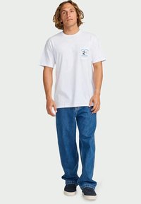 Hombre con el cabello hasta los hombros vestido con una camiseta gráfica blanca, jeans azules y zapatillas negras, de pie contra un fondo liso.