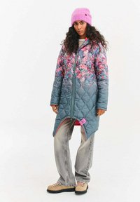 Prošívaná bunda s kvetinovým vzorom, prechod z ružovej do modrej. Má zapínanie na zips, bočné rozparky a kontrastnú ružovú podšívku. Neformálne šedé džínsy a čižmy.