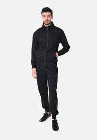 Giacca nera con zip e pantaloni coordinati realizzati in tessuto morbido. Presenta polsini e colletto a costine, abbinati a sneakers nere.