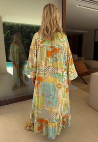 Blonds cilvēks, valkājot garu, krāsainu rakstainu kleitu, stāv telpās, raugoties uz stikla durvīm, kas atspoguļo viņu priekšpusi, blakus atrodas mūsdienu mēbeles.