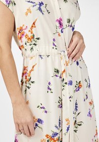 Bloemenjurk van crèmekleurige stof, met veelkleurige bloemen (paars, oranje, roze) en een gestrikt taille-detail; knoopsluiting aan de voorkant.