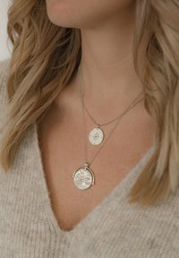 Ezüst rétegezett nyakláncok, egy kör alakú medállal, amelyen gravírozások találhatók, és egy kisebb ovális medállal, amely csillagmintás. Texturált szövet háttér.