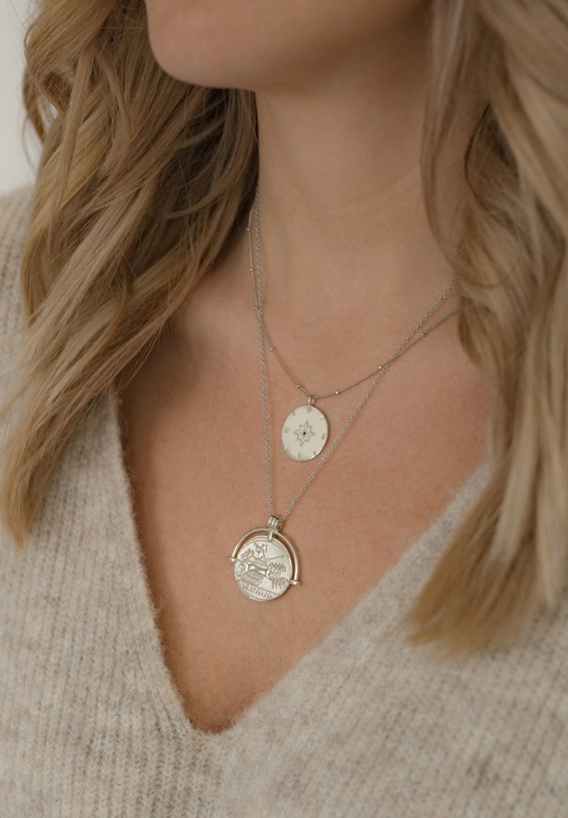 Ezüst rétegezett nyakláncok, egy kör alakú medállal, amelyen gravírozások találhatók, és egy kisebb ovális medállal, amely csillagmintás. Texturált szövet háttér.