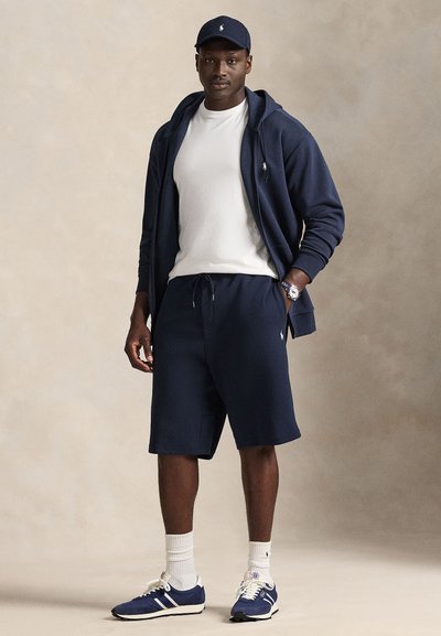 Φούτερ με κουκούλα και σορτς σε ναυτικό μπλε χρώμα από βαμβακερό μείγμα, συνδυασμένα με λευκό T-shirt. Αξεσουάρ περιλαμβάνουν ένα ναυτικό καπέλο και αθλητικά παπούτσια με ρίγες.