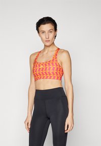 Sportbehå i klar orange med gula bladmönster, med en sömlös design och breda axelremmar. Kombinerad med svarta högmidjade leggings.