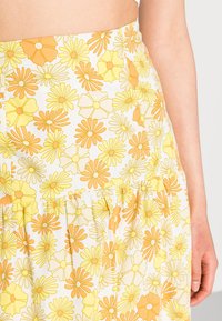 Saia de estampado floral em tons de amarelo e laranja, apresentando um design em camadas e um tecido leve com uma textura suave. Padrões florais por toda a parte.