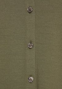 Camisa verde de botones con una tela texturizada y cuatro botones redondos, dos de los cuales tienen un contraste de color más oscuro.