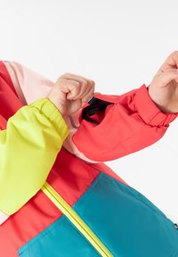 Färgglad jacka med röda, rosa och akvafärger i sektioner, med elastiska manschetter och en ljusgul detalj på ärmarna. Inkluderar en ficka med dragkedja.