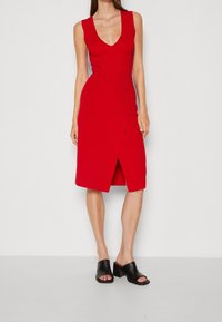Robe rouge sans manches avec un col en V et un design portefeuille, longueur genoux avec une fente. Portée avec des chaussures noires à talons blocs ouverts à l'avant.
