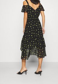 Frau, die ein schwarzes, transparentes Midikleid mit grünem Blumenmuster und kalten Schultern trägt, kombiniert mit schwarzen Absatzsandalen, mit dem Rücken zugewandt.