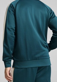 Tyrkysová sportovní mikina s texturovanou tkaninou, kontrastními béžovými bočními panely a žebrovanými manžetami. Jednoduchý design s uvolněným střihem.