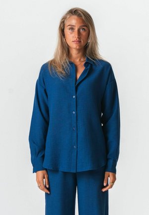 Vrouw met lang blond haar, gekleed in een losse blauwe blouse met knopen en bijpassende blauwe broek, staand tegen een effen lichte achtergrond.