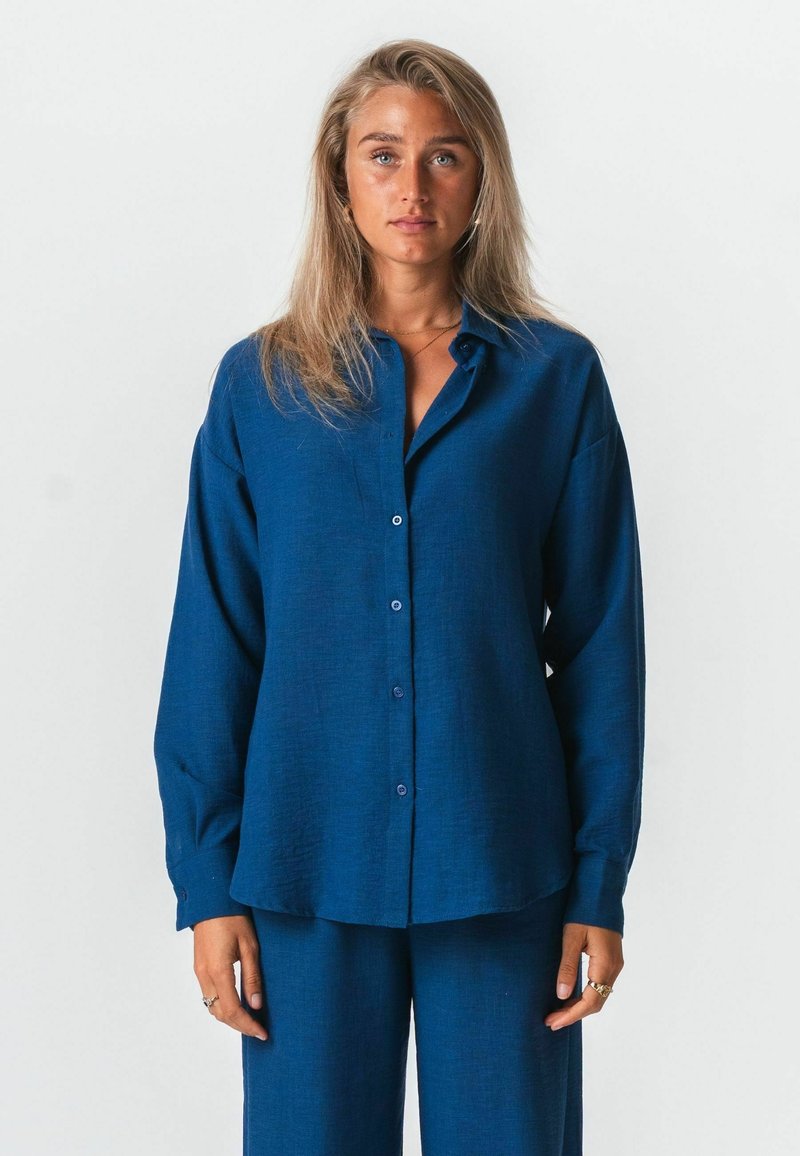 Femme aux longs cheveux blonds portant une chemise bleue ample à boutons et un pantalon bleu assorti, debout devant un fond clair uni.
