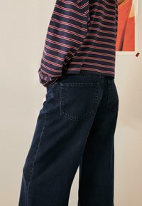 Sötét farmernadrág széles szárú kialakítással, két hátul zsebbel. Egy tengerészkék-fehér csíkos hosszú ujjú felsővel párosítva.