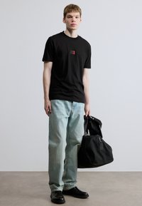 Μαύρο βαμβακερό μπλουζάκι με μια μικρή κόκκινη ετικέτα, ανοιχτό μπλε τζιν με ίσιο κόψιμο και μαύρα δερμάτινα μποτάκια. Κρατάει μια μαύρη τσάντα duffel.