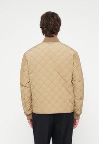 Béžová prešívaná bomber bunda s rebrovaným golierom a lemy na rukávoch, má textúrovaný povrch a voľný strih. Viditeľné švy bez kovových doplnkov.