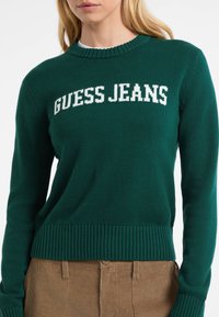 Zeleno pleteno pulover s rebrastimi manšetami in spodnjim delom. "GUESS JEANS" v belem tisku čez prsa. Tekstura tkanine in krojen dizajn s krožnim izrezom.