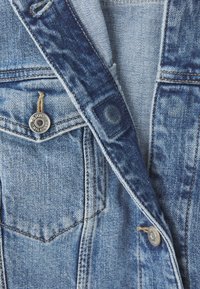 Denim jakk, mis sisaldab heledaid ja tumedaid siniseid toone, kulunud tekstuuri ning metallist riistvara, millel on nuppude peal logo.