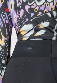 Kadar izbliza osobe koja nosi gornji dio s više boja i uzoraka te crne tajice visokog struka s diskretnim Adidas logom na pojasu.