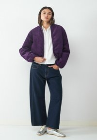 Kobieta w fioletowej kurtce bomberce z korduroy, białej koszuli, szerokich ciemnych dżinsach i srebrnych sneakersach, stoi na tle białego tła.