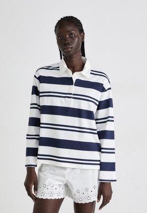 Langermet polo i marineblå og hvite striper, med en kontrastfarget hvit krage og knappestolpe, sammen med hvite shorts med hullmønster.