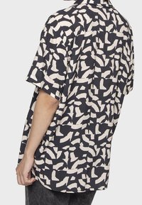 Camicia a maniche corte con colletto in tessuto blu navy, decorata con motivi astratti bianchi; vestibilità rilassata, texture liscia e orlo curvo.