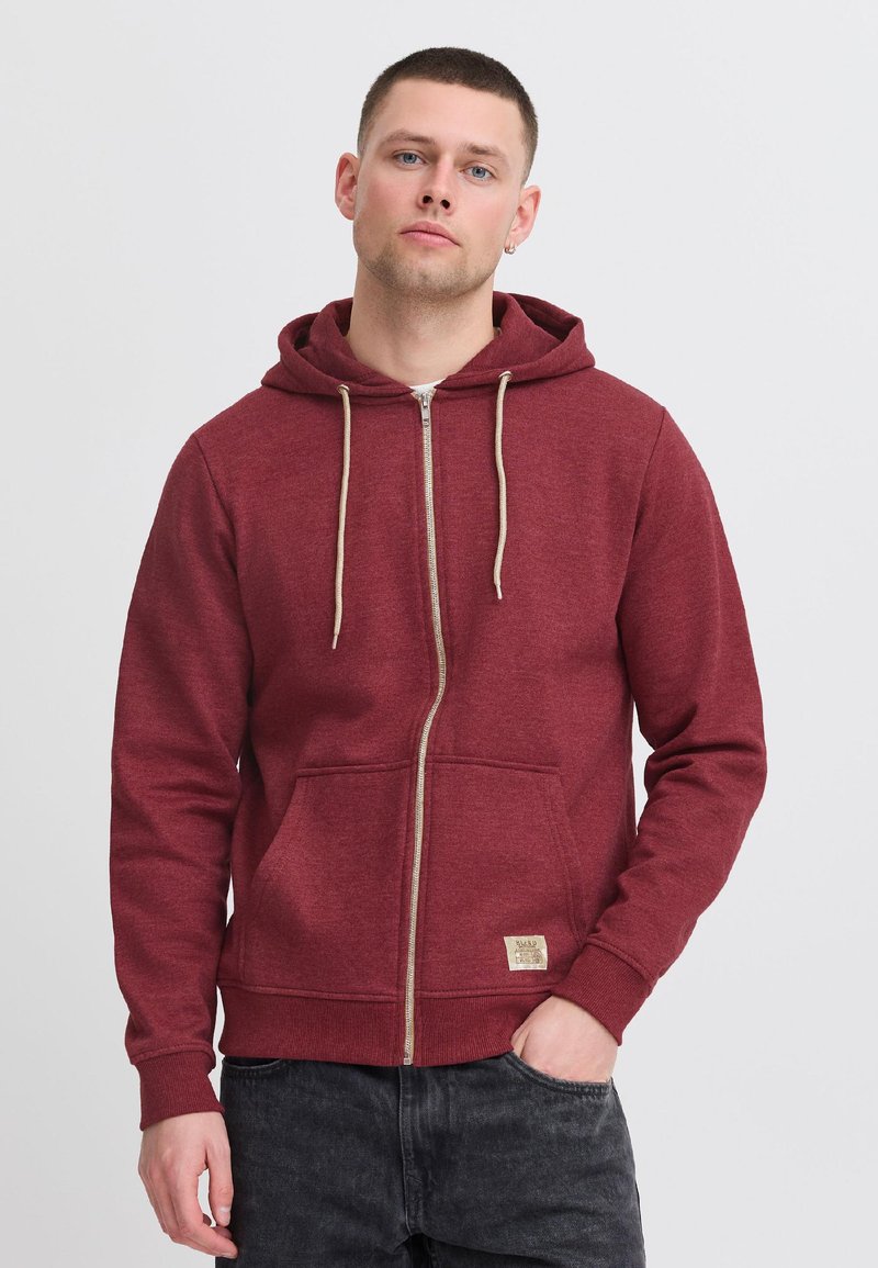 Jonge man draagt een bordeauxrode hoodie met rits, beige trekkoorden en zwarte spijkerbroek, staand met één hand in de zak tegen een effen achtergrond.