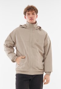 Giacca beige chiaro con zip frontale, cappuccio, polsini elasticizzati e due tasche laterali. Realizzata in un materiale leggero e texturizzato.