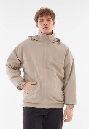 Veste beige clair avec zip sur le devant, capuche, poignets élastiques et deux poches latérales. Fabriquée dans un matériau texturé et léger.
