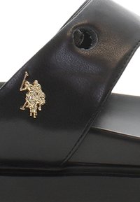 Musta nahkasaapas, jossa on kirkas kultainen logoyksitys. Sileä pinta, minimaalinen muotoilu ja avoin varvas.