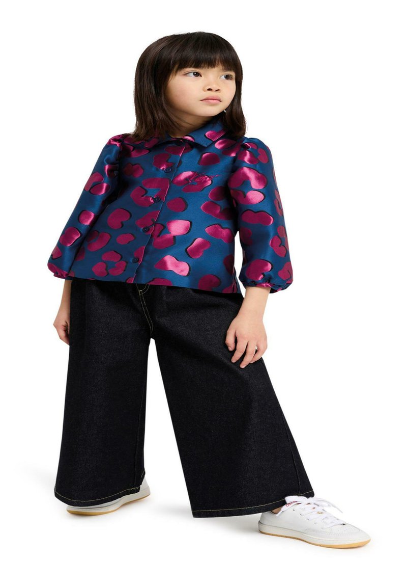 Blauwe satijnen blouse met een roze abstract patroon, korte pofmouwen, gecombineerd met wijde donkere spijkerbroek en witte sneakers.