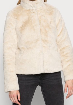 Person mit einer hellbeigen Kunstpelzjacke mit hohem Kragen und langen Ärmeln, kombiniert mit einer schwarzen Hose, von den Schultern bis zur Hüfte gezeigt.