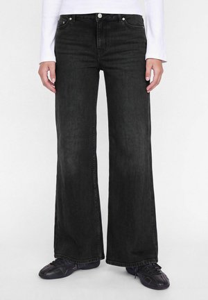 Une personne porte un jean noir à jambes larges avec poches avant, un haut blanc et des baskets noires sur un fond uni.