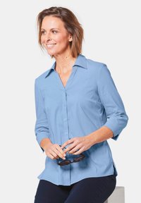 Camicia blu chiaro con colletto a punta, maniche a tre quarti e spacco laterale, abbinata a pantaloni scuri e occhiali da sole.