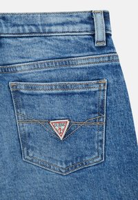 Achterzak van blauwe spijkerbroek met oranje stiksels en een driehoekige, witte en rode Guess-merkpatch.