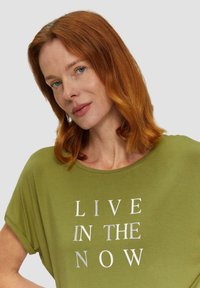 Olijfgroene, losse t-shirt met ronde halslijn en korte mouwen. Voorzien van "LIVE IN THE NOW" in zilveren tekst op de voorkant.