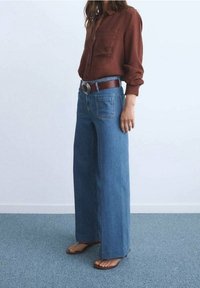 Chemise marron à manches longues avec poche avant, associée à un jean en denim bleu à jambes larges. Ceinture marron avec une boucle argentée. Sandales plates.