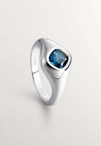 Anillo de plata con un diseño suave y curvado, que presenta una piedra azul facetada engastada en un bisel asimétrico. Acabado pulido, forma moderna.