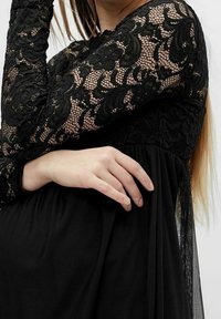 Svart klänning med långa spetsärmar och en mjuk tygkropp. Har intrikata spetsmönster och en flödande kjol med veck.