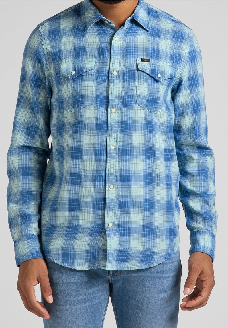 Homme portant une chemise à carreaux bleue et vert clair avec poches poitrine et jean bleu, visible du cou aux hanches.
