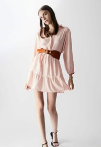 Rosa Satinhemdkleid mit langen Ärmeln, Knopfleiste und gerafftem Saum. Akzentuiert mit einem braunen Gürtel. Getragen mit schwarzen Riemchensandalen.