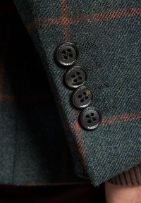 Wełniana marynarka w niebiesko-zielony wzór w jodełkę, z czterema czarnymi guzikami o lekko połyskującej powierzchni przy mankietach rękawów.
